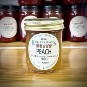 Peach jam in an 8 oz jar from King Orchards, labeled family grown with peaches, sugar, lemon juice, and pectin, on a farm market shelf beside jars of Montmorency cherry jam