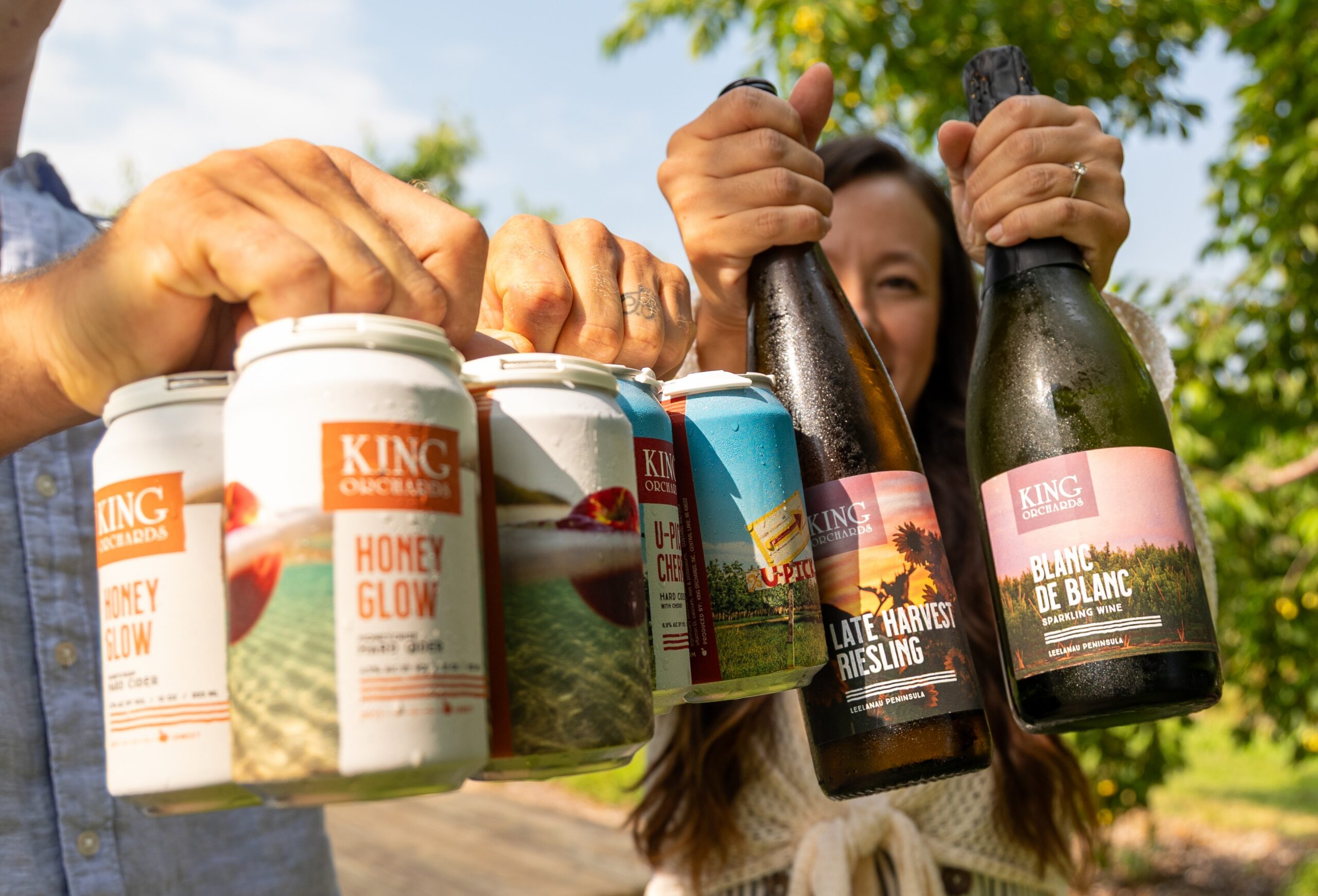 two people holding hard cider cans and wine bottles in the orchard